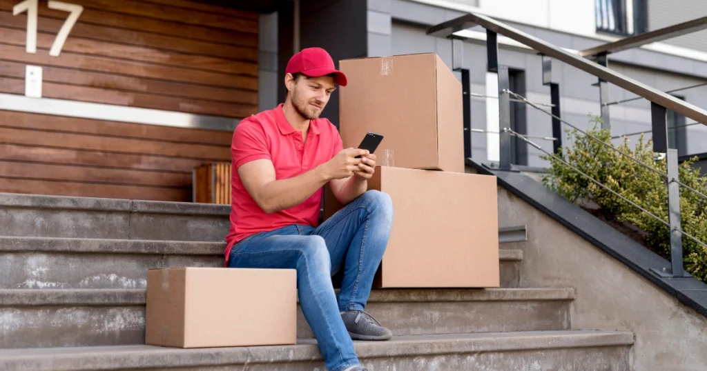 A product delivery service worker in a red uniform and cap sits on outdoor steps next to several cardboard boxes, checking his phone. The background features a modern residential building with the number 17 displayed.