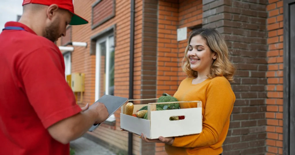 A product delivery service worker in a red uniform uses a tablet while delivering a crate of fresh vegetables to a smiling woman at her doorstep. A brick residential building is visible in the background.