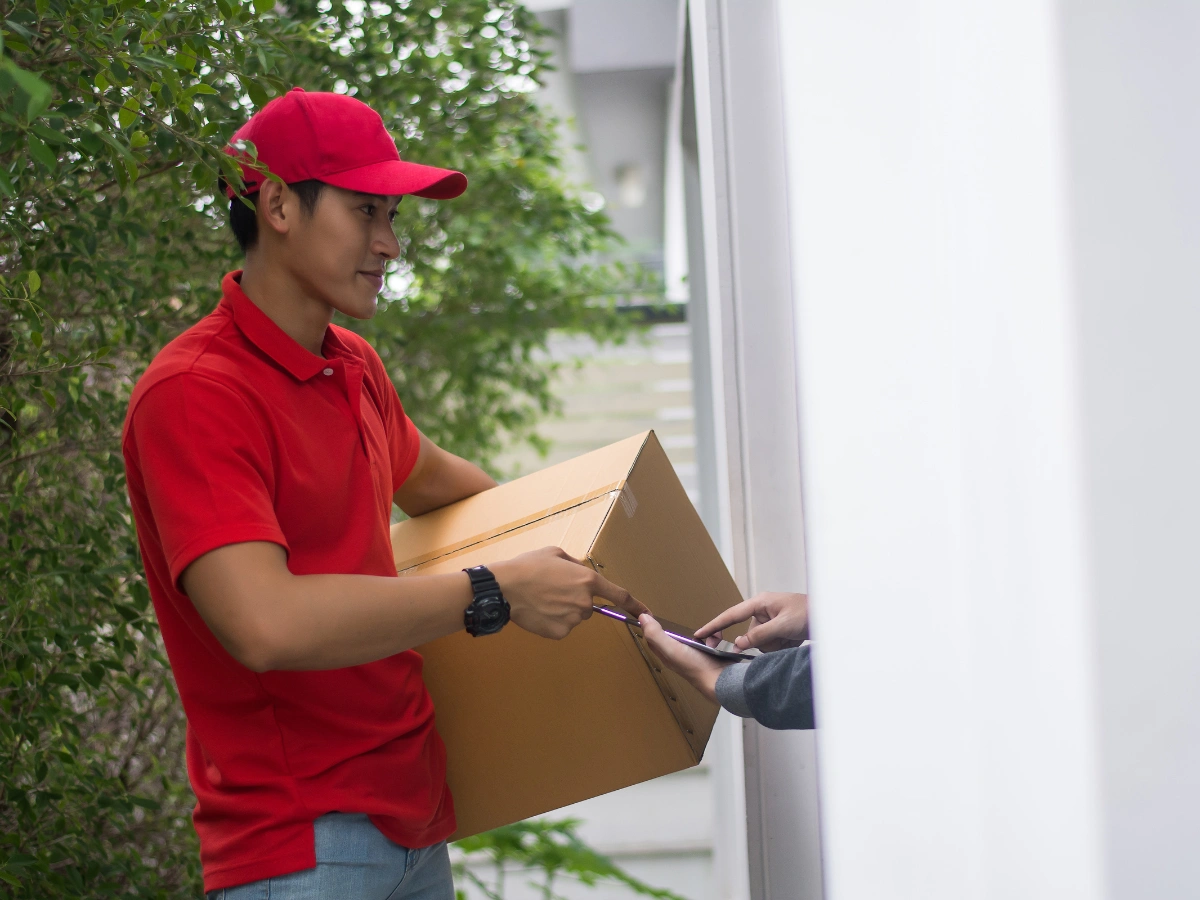 A product delivery service worker in a red shirt and cap delivers a large cardboard box to a customer at their doorstep. The customer signs for the package on a handheld device.