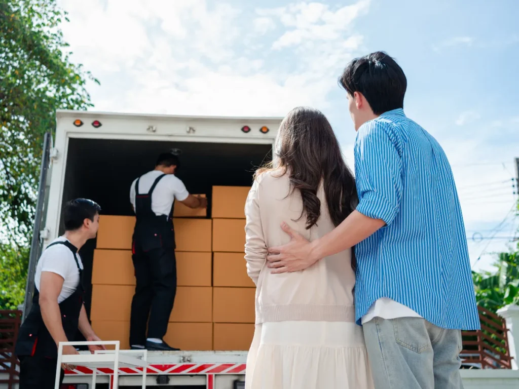 A couple watches as two removal services workers load boxes into a truck. The workers in overalls organize boxes. Clear sky outdoors.