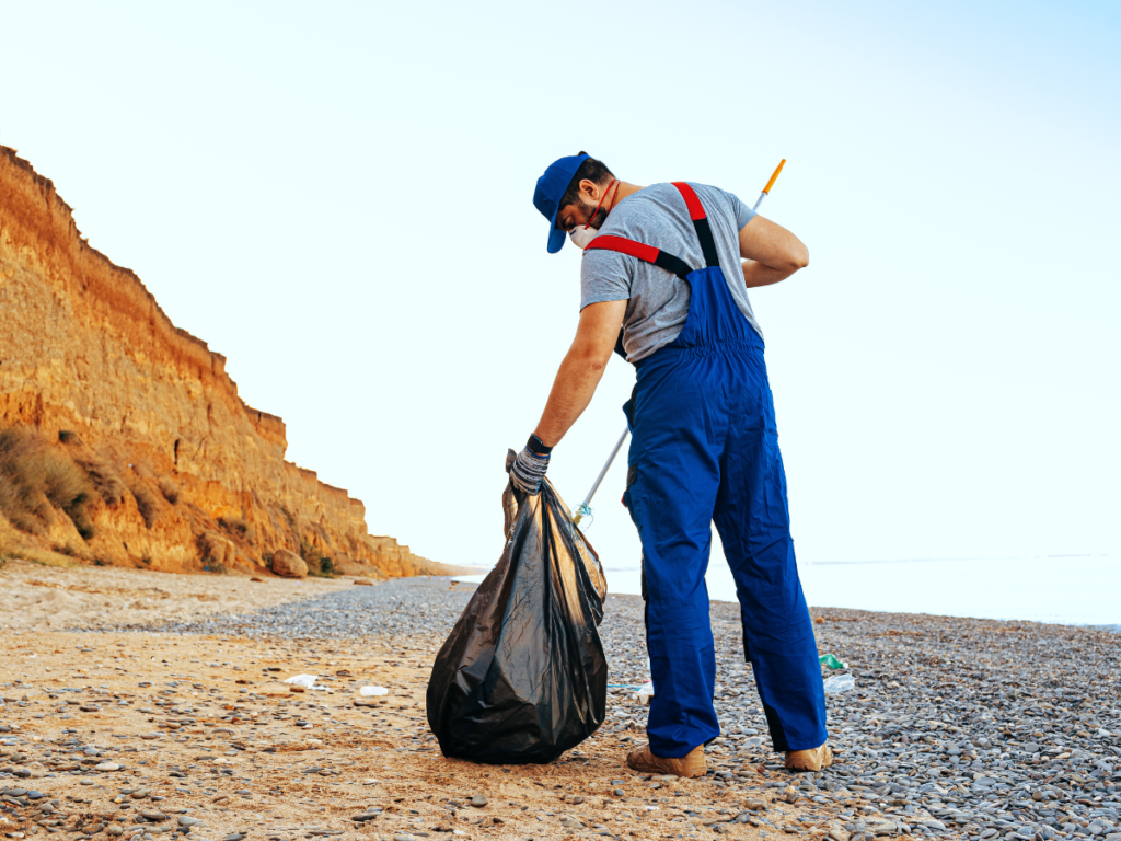 Worker collecting trash in a black bag on a rocky beach as part of waste removal services.